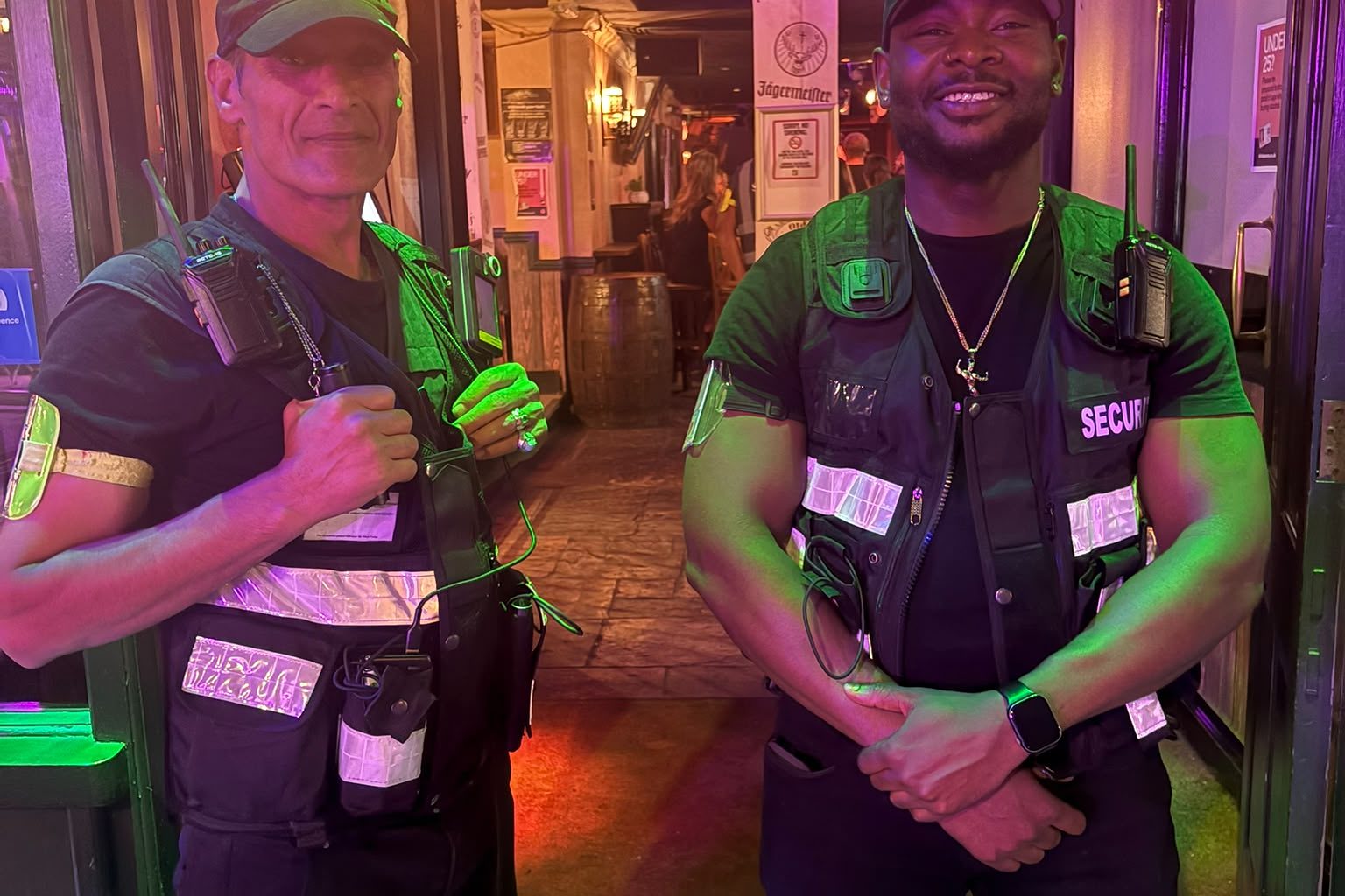 Two door supervisors on duty at a venue entrance under green lighting at night.